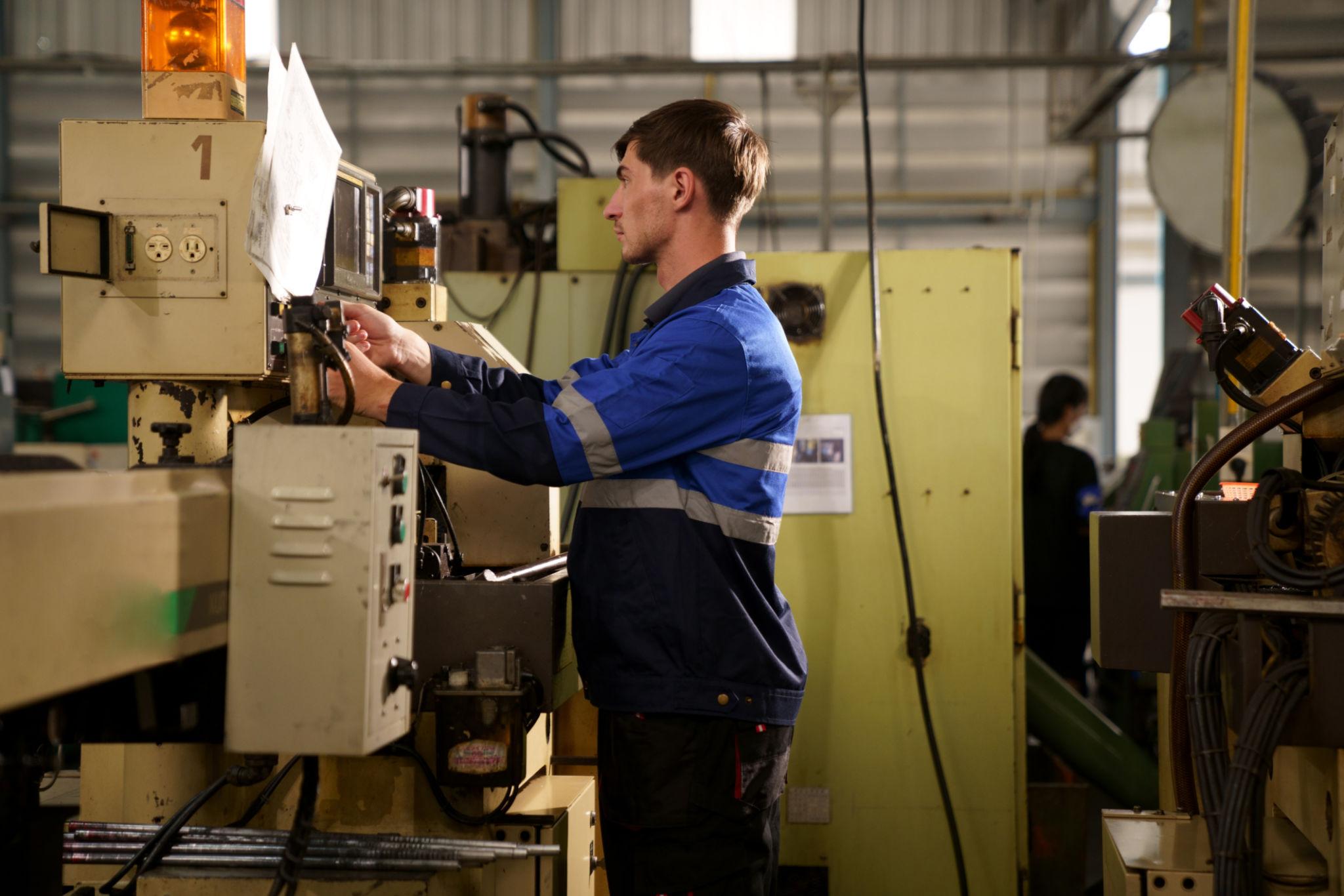 A machinist in a blue work jacket operating a control panel on industrial factory machinery.
