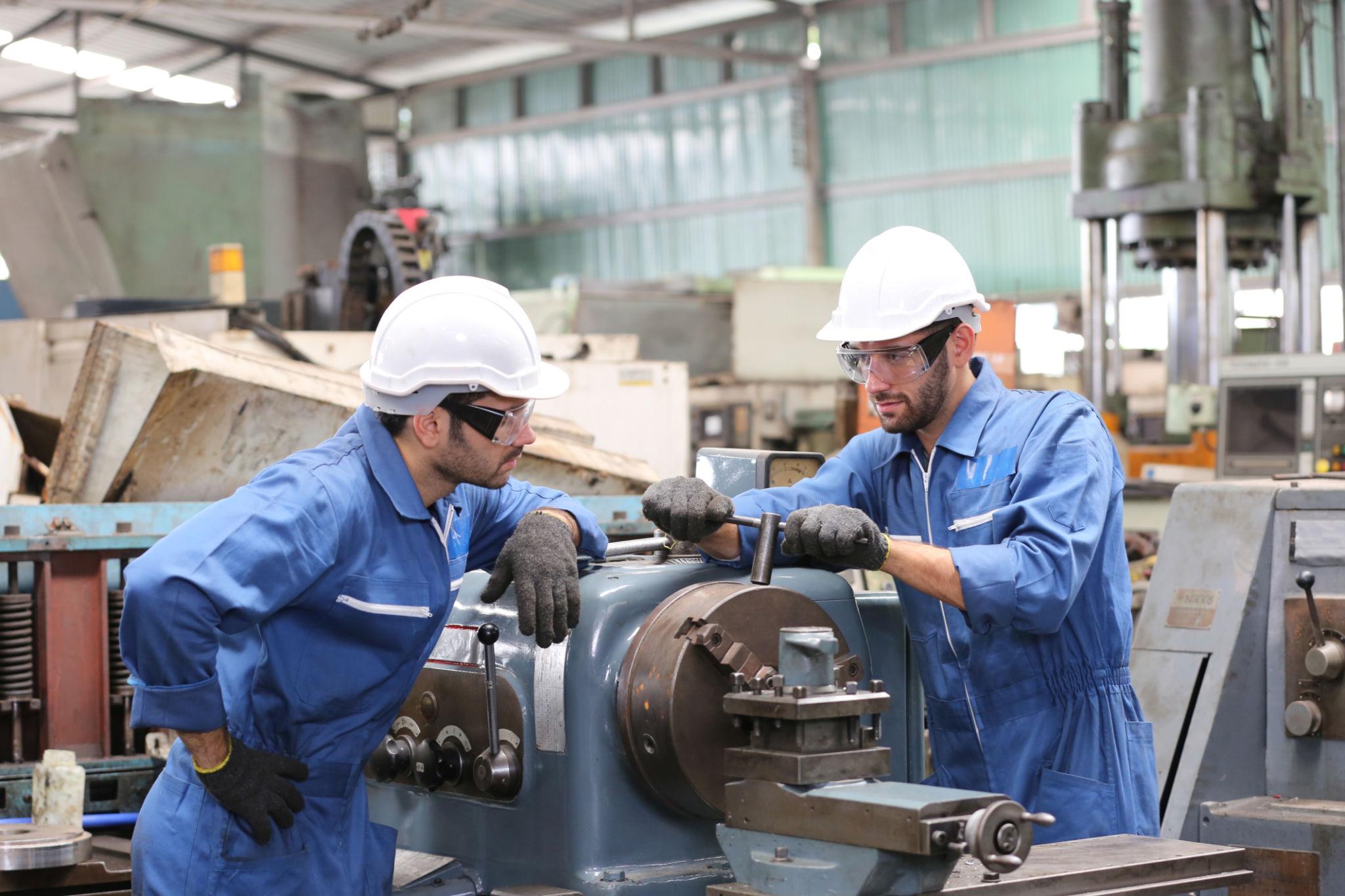 Two factory workers in blue jumpsuits and hard hats operating heavy machinery shop equipment.